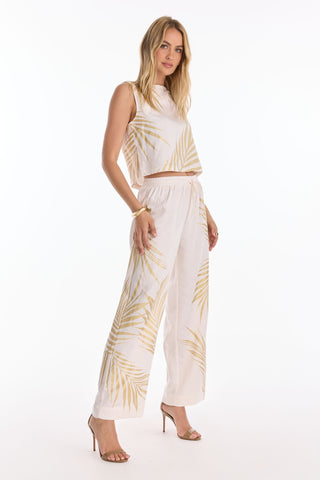 A woman wears the Sola Linen Pant - Gold Palm, featuring wide-leg styling and a light beige palm leaf print on white. Paired with nude heels, set against a white background. Preorder now; ships June 25.