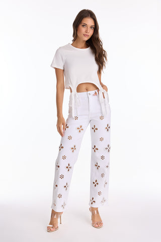 A woman confidently poses in a white Diana Satin Bow Shirt paired with high-waisted floral-cutout white pants and open-toe heels, set against a plain white background.