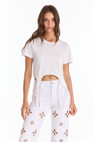 A woman wears a Diana Satin Bow Shirt - White, a short-sleeve white crop top with tie details at the hem, paired with high-waisted jeans with brown and tan floral cutouts. She stands against a plain white background.
