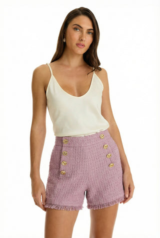 A woman with long brown hair wears a white sleeveless top and the Tamara Tweed Button Short - Blush, featuring gold buttons, fringe details, and a gold side zipper, standing against a plain white background.