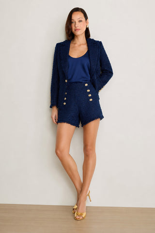A woman stands against a light wall wearing the Tamara Tweed Button Short in navy, paired with a matching textured blazer, a silky blue top, and gold high-heeled shoes.