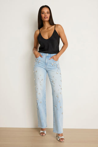 A woman with straight hair stands by a plain wall, hands in her pockets, wearing a black sleeveless top and the Tara Crystal Jean - Light Blue paired with silver open-toe heels.