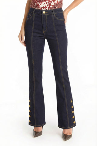 Wearing the Veronica Button Jean in Indigo—high-waisted dark blue flare jeans with gold outer seam buttons, four-way stretch denim, and a leg-lengthening fit—paired with a red and white patterned top and pointed-toe heels against a plain white background.