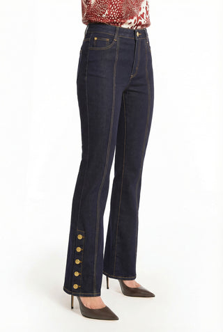 Wearing the Veronica Button Jean - Indigo, featuring gold buttons along the lower outer seam, and brown pointed-toe heels, a person stands against a white background, creating a flattering, leg-elongating silhouette.