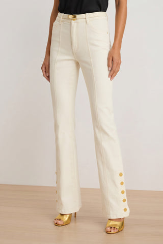 A woman stands on a wooden floor against a plain background, wearing the Veronica Button Jean in ivory with gold buttons at the ankles, gold open-toe heels, and a black top.