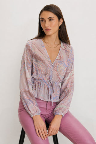 A woman with long brown hair sits on a stool against a light background, wearing the Whitney Chiffon Top – Pastel Paisley, a feminine blouse with a delicate button front and drawstring waist, paired with high-waisted mauve pants.