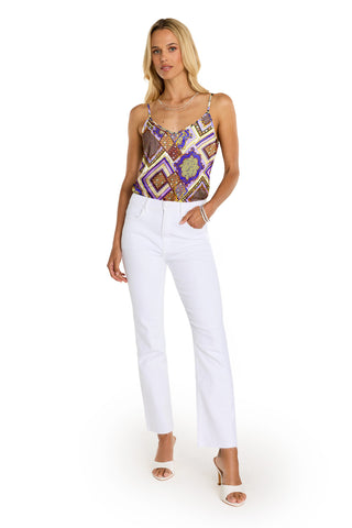 A woman stands against a white background, wearing the Yara Silk Camisole - Riviera Chain in purple and brown tones, paired with white straight-leg pants and white open-toe heels—an effortlessly chic resort look.