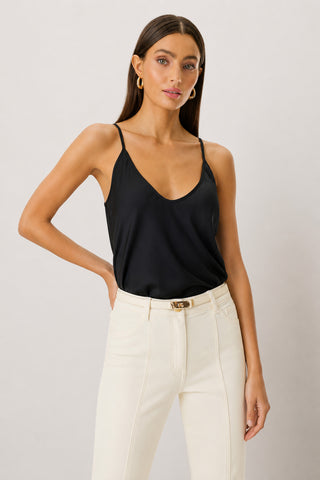 A woman with long brown hair wears the Yara Silk Camisole in black, featuring adjustable straps, paired with high-waisted white pants. She stands with one hand on her hip against a plain light background.