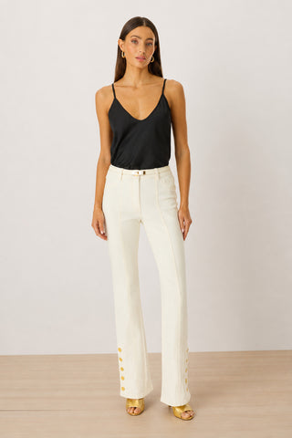 A woman faces forward in the Yara Silk Camisole - Black with adjustable straps, high-waisted white pants with gold buttoned cuffs, gold hoop earrings, and gold open-toe heels against a plain light background.