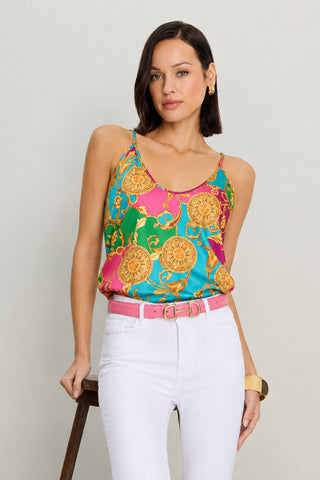 A woman with straight dark hair wears the Yara Silk Camisole in Dolce Sun, styled with white high-waisted pants and a pink belt. She stands against a neutral background, resting one hand on a stool.