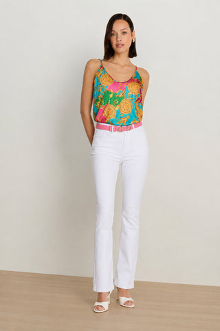 A woman with short brown hair poses relaxed against a plain background, wearing the Yara Silk Camisole - Dolce Sun—a colorful piece with a pink and green Mediterranean print—paired with white high-waisted pants, a pink belt, and white heeled sandals.