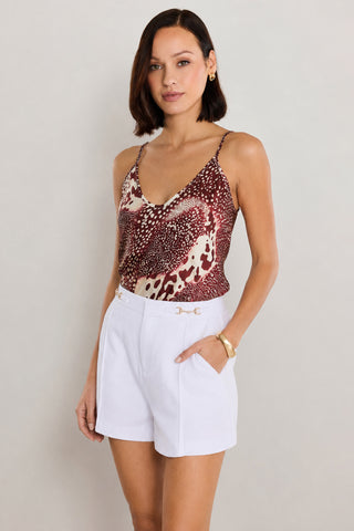A woman wearing the Yara Silk Camisole in Garnet Crush—a sleeveless, red-and-white patterned top—paired with white high-waisted shorts with pockets, poses against a plain light background. She has chin-length dark hair and gold jewelry.