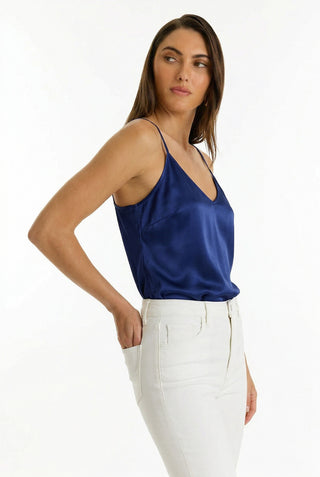 A woman with long brown hair wears the Yara Silk Camisole - Navy, featuring a deep v-neck, paired with high-waisted white pants. She stands against a plain white background, looking left with one hand in her pocket.