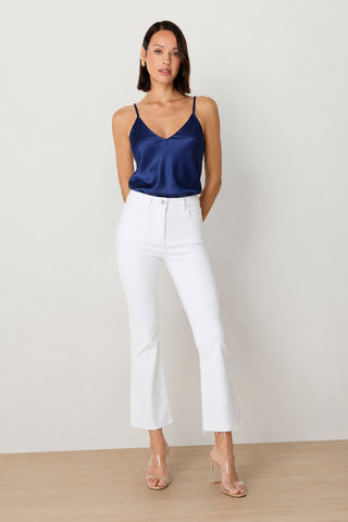 A woman stands against a plain background wearing the Yara Silk Camisole in navy with adjustable straps, high-waisted white jeans with frayed hems, and clear heeled sandals. Her hair is straight and she has one hand in her pocket.
