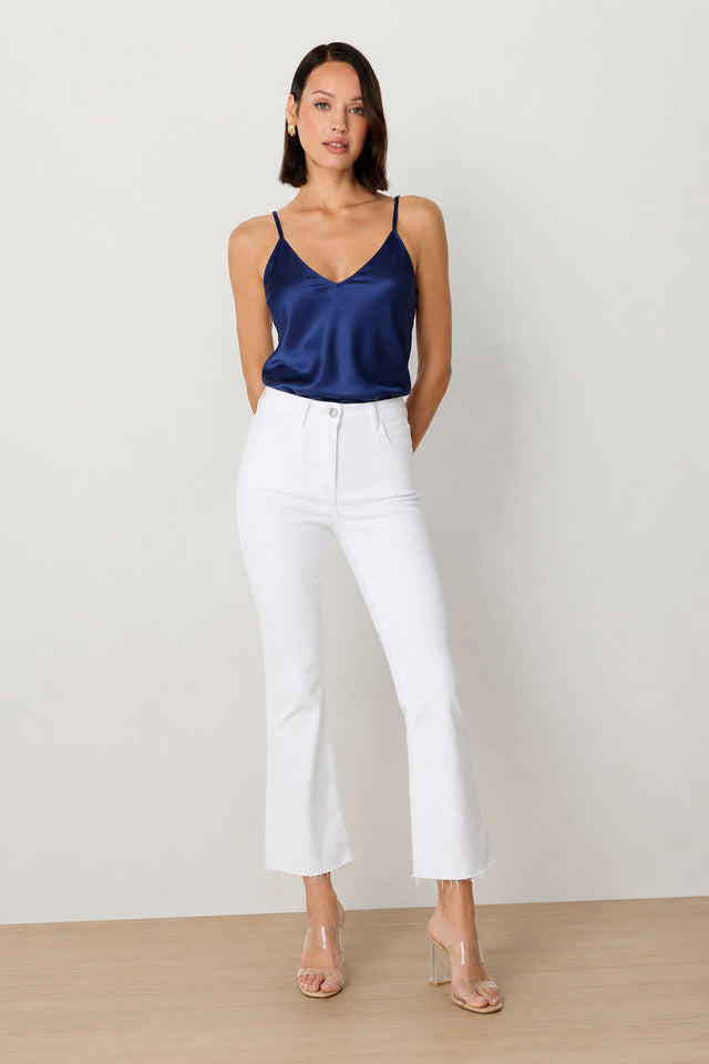 A woman stands against a plain background wearing the Yara Silk Camisole in navy with adjustable straps, high-waisted white jeans with frayed hems, and clear heeled sandals. Her hair is straight and she has one hand in her pocket.