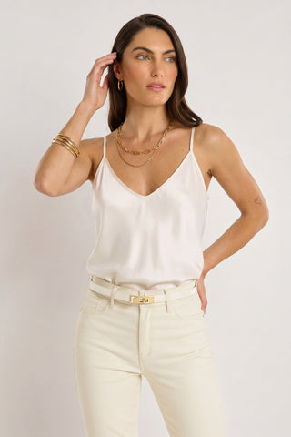 A woman with long brown hair, gold jewelry, and a small heart tattoo on her left arm models the Yara Silk Camisole - Pearl with adjustable straps and cream pants against a plain background.