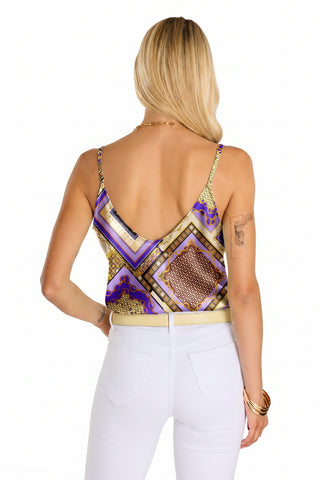 A woman with long blonde hair stands facing away, wearing the Yara Silk Camisole - Riviera Chain in a patterned purple, gold, and brown design with adjustable straps, paired with fitted white pants, gold jewelry, and a tattoo on her upper left arm.