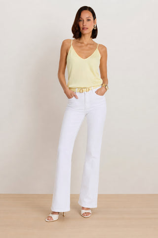 A woman stands on a wooden floor against a plain background, hands in her pockets, wearing the Yara Silk Camisole in Sunshine Yellow, paired with white high-waisted pants, white heeled sandals, gold earrings, a gold bracelet, and a gold belt.