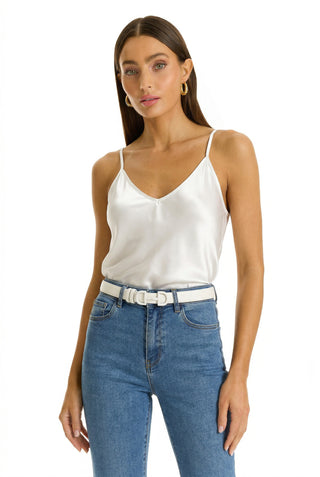 A woman wears the Yara Silk Camisole in white with adjustable straps, high-waisted blue jeans, a white belt, and gold hoop earrings, standing against a plain white background.
