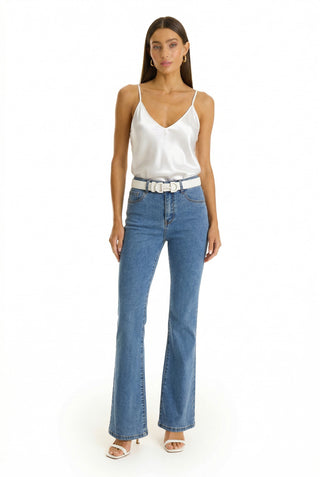 A woman stands against a white background, wearing a white Yara Silk Camisole with adjustable straps, blue high-waisted flared jeans, a white belt, and white heeled sandals. She has long brown hair and neutral makeup.