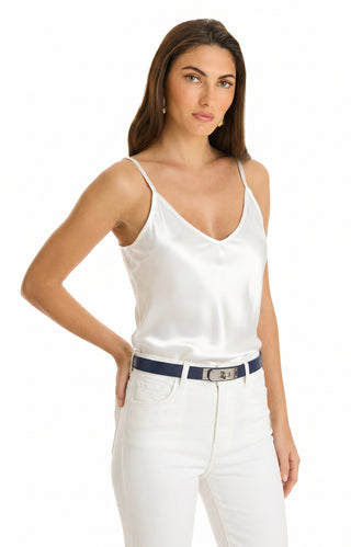 A woman with long brown hair wears the Yara Silk Camisole in white, paired with white jeans and a navy blue belt. She stands confidently, hand on hip, against a plain white background.