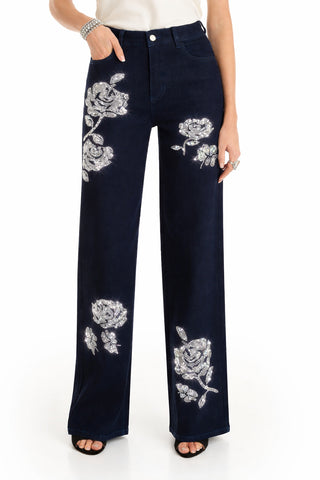 The lower half of a person is shown wearing the Kate Crystal Rose Jean in Indigo—high-waisted, wide-leg denim pants with large silver floral embroidery—paired with black open-toe shoes and a white top.